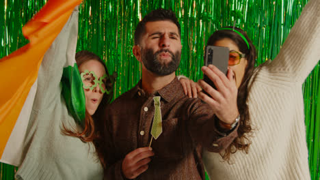 Studio-Shot-Of-Friends-Dressing-Up-With-Irish-Novelties-And-Props-Posing-For-Selfie-Celebrating-St-Patrick's-Day-Against-Green-Tinsel-Background-1
